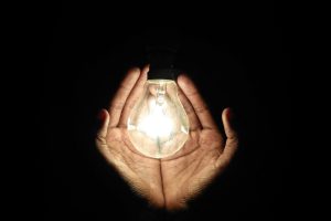 A pair of hands gently cradles a glowing light bulb, illuminating the darkness.